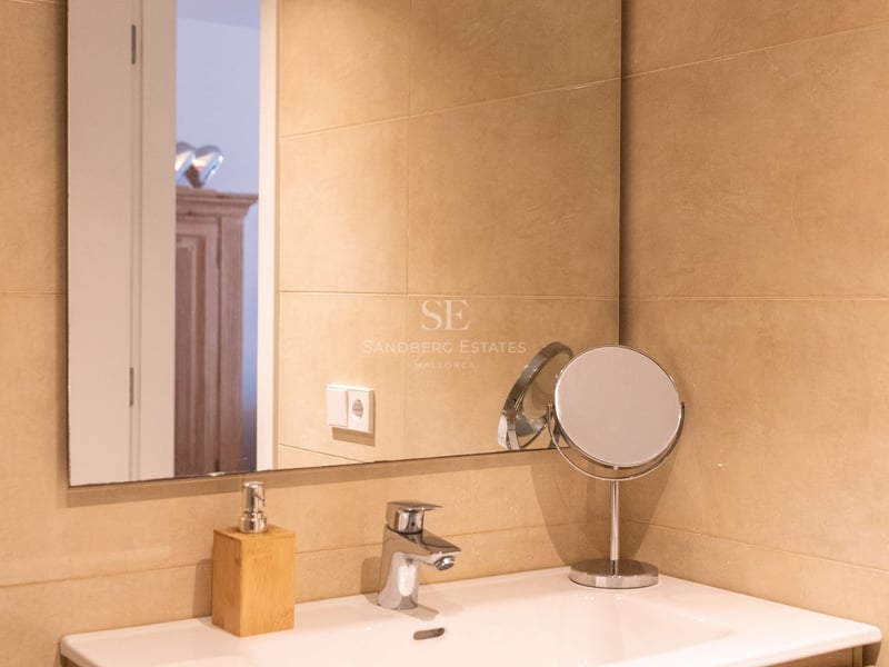 Weißes Keramikwaschbecken auf hellem Holzunterschrank, großer Spiegel und sandfarbene Wandfliesen.