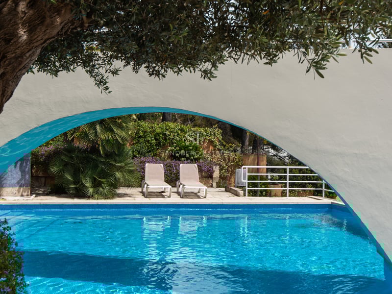 Blue swimming pool and sun loungers framed by a white architectural arch and lush Mediterranean greenery.
