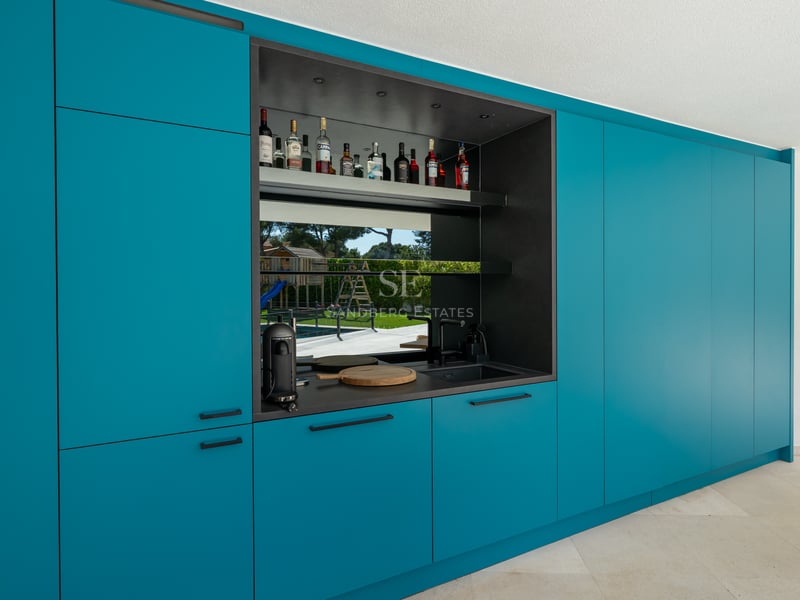 Mobilier de cuisine moderne bleu canard avec bar intégré et reflet du jardin dans la crédence en verre.