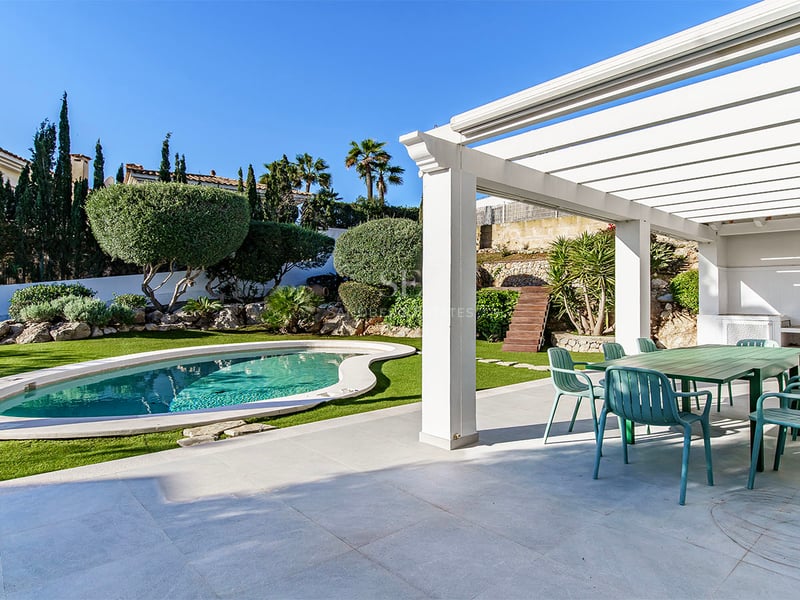 Vit pergola som täcker en matplats utomhus med gröna stolar bredvid en njurformad pool och prunkande trädgård.