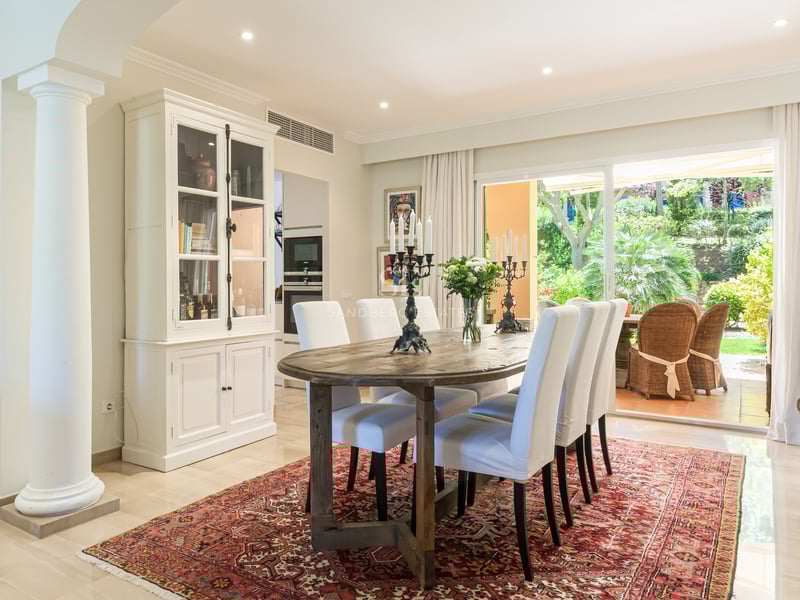 Dining room featuring an oval wooden table, white upholstered chairs, and an oriental rug, overlooking a lush garden.
