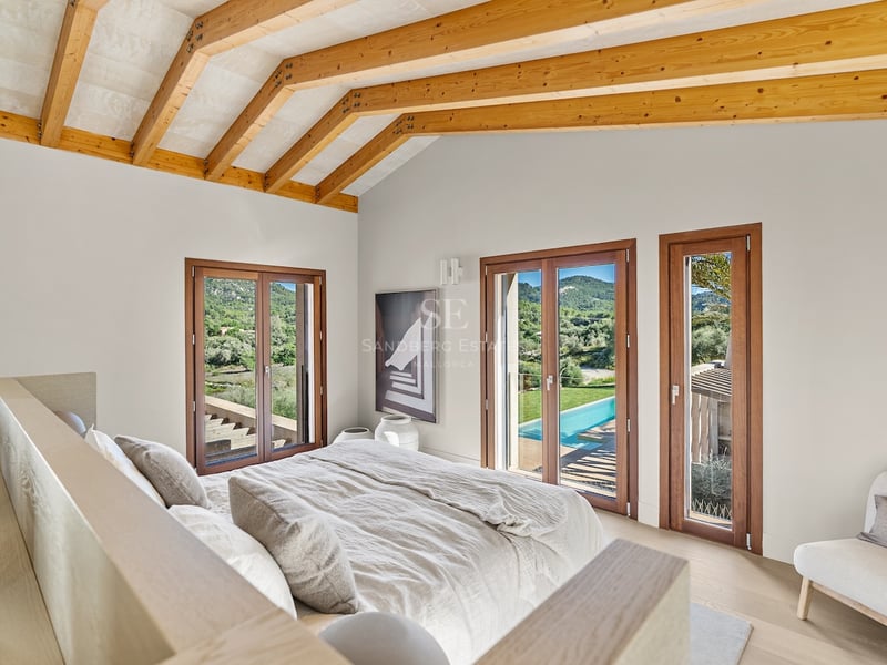 Chambre moderne avec plafond à poutres en bois, lit en lin beige et portes vitrées donnant sur une piscine et collines.