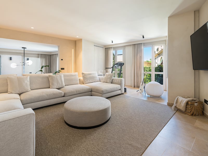 Un salon moderne et lumineux avec un grand canapé d'angle crème, un pouf rond et des sols en pierre naturelle.