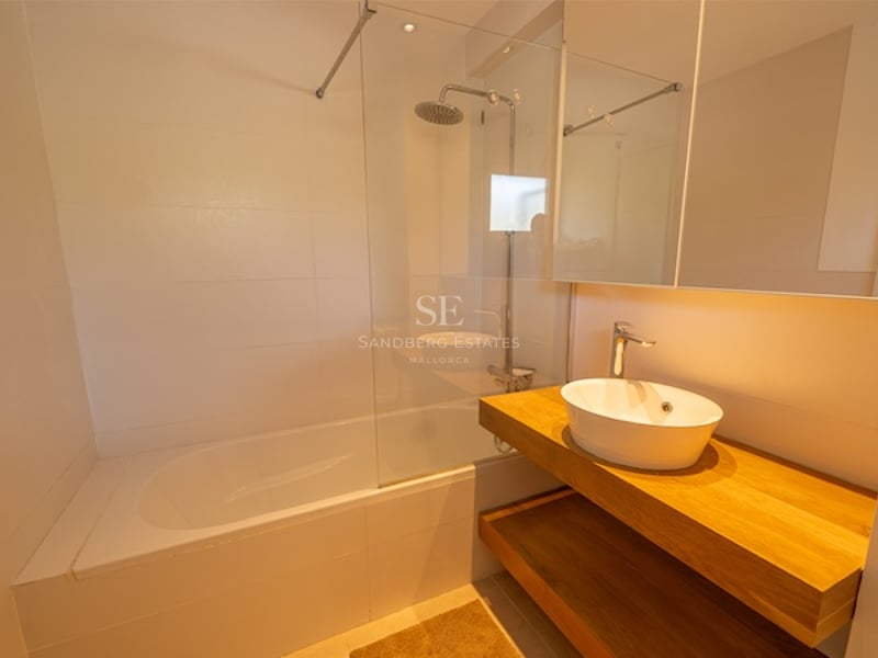 Baño minimalista con bañera blanca, mampara de cristal, ducha de efecto lluvia y mueble de madera con lavabo sobre encimera.