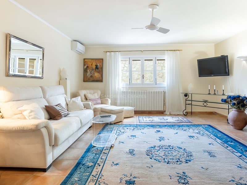 Salon lumineux avec canapé blanc, grand tapis bleu à motifs, sol en terre cuite et ventilateur de plafond.