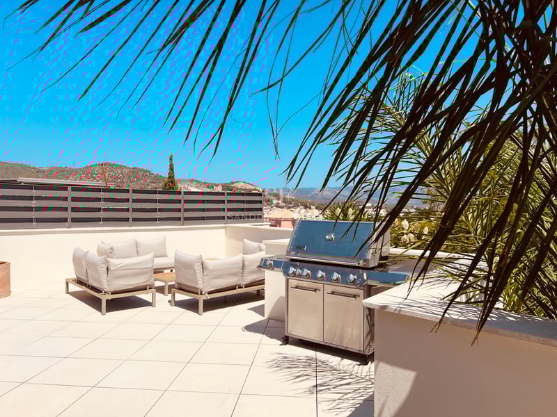 Toit-terrasse ensoleillé avec grand barbecue en inox, salon de jardin moderne et vue sur les montagnes.