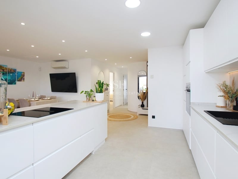 Ljust vitt modernt kök med köksö, svart diskho och infälld belysning mot vardagsrummet.