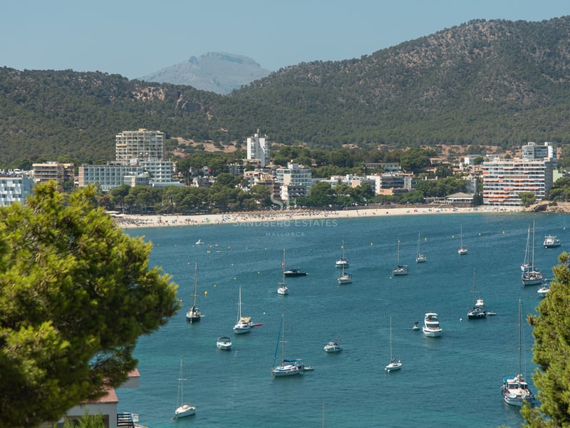 Vue surélevée d'une baie turquoise avec des voiliers, une plage de sable et des montagnes vertes sous un ciel dégagé.