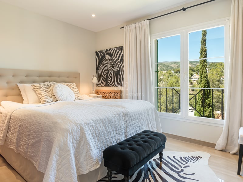 Bright bedroom featuring a large bed with a tufted headboard and a window overlooking lush green hills and a cypress tree.