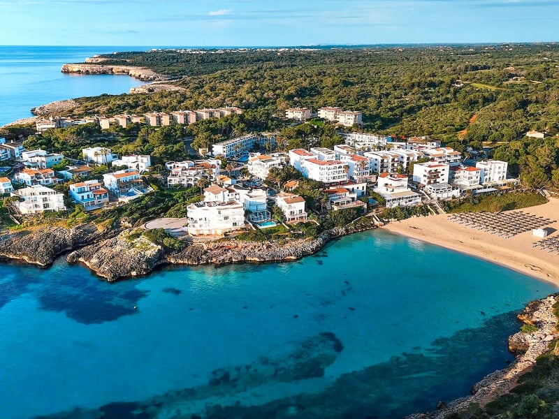 Vue aérienne d'un complexe de bâtiments blancs à côté d'une crique turquoise et d'une plage entourée d'une forêt verte.