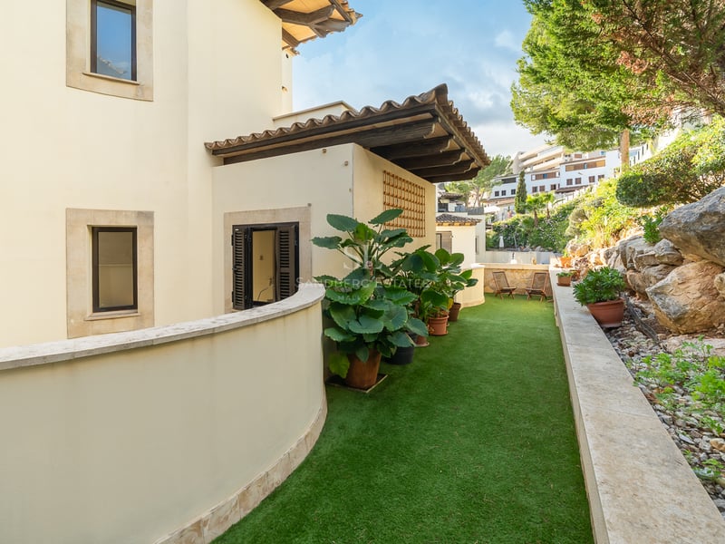 Böjd krämvit vägg bredvid en gångväg med konstgräs, kantad av ett stenparti och krukväxter.