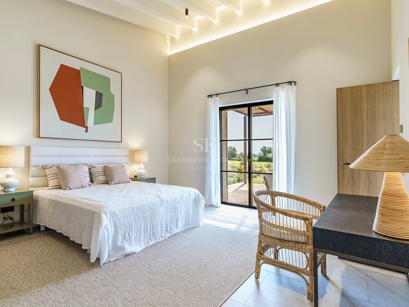 Helles, modernes Schlafzimmer mit hohen Decken, weißem Bett, Korbstuhl und großen Glastüren zum Garten.