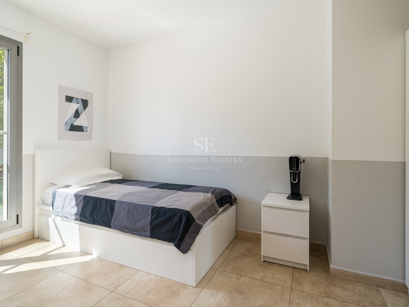 Ett ljust sovrum med en vit enkelsäng, klinkergolv, vit garderob och ett stort fönster med utsikt över träd.