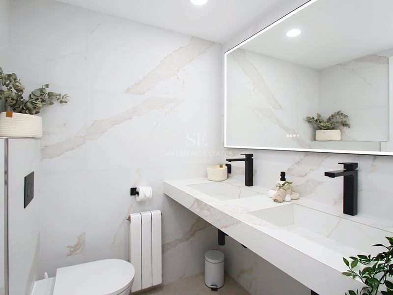 Modernt badrum med vita marmorliknande väggar, dubbeltvättställ med svarta kranar och en stor bakgrundsbelyst spegel.