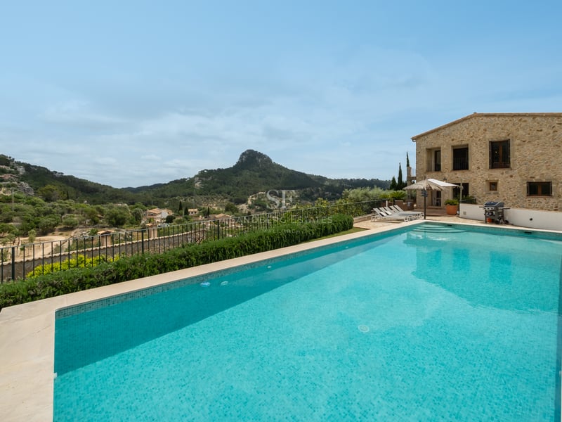 Grande piscine rectangulaire surplombant une vallée verdoyante et des montagnes avec une villa en pierre.