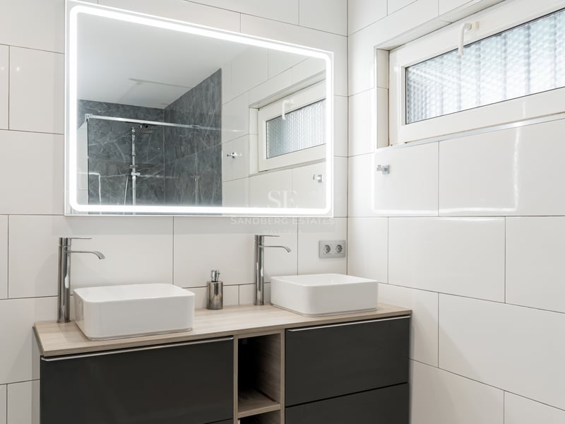 Salle de bain moderne avec deux vasques carrées, un grand miroir éclairé et des murs en carrelage blanc.
