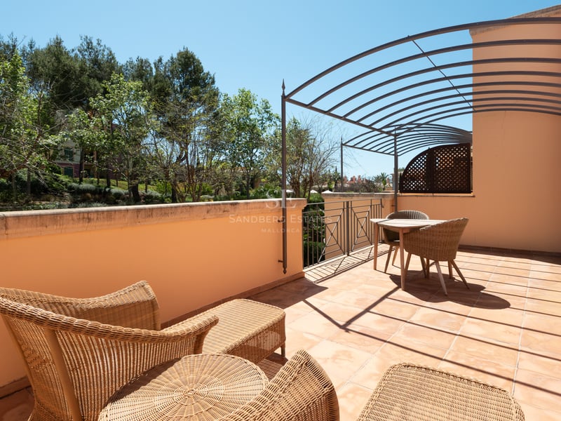 Spacious terrace featuring terracotta tiles, wicker furniture, and a metal pergola overlooking lush trees.