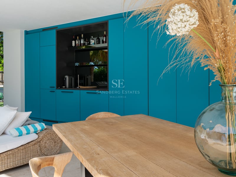 Terrasse avec armoires bleu sarcelle, station bar, table en bois clair et méridienne en osier.