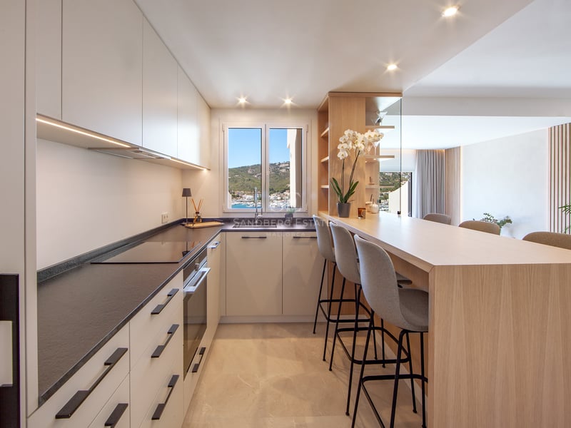 Cuisine moderne avec placards blancs, plans de travail sombres, bar en bois avec tabourets et fenêtre avec vue sur le port.