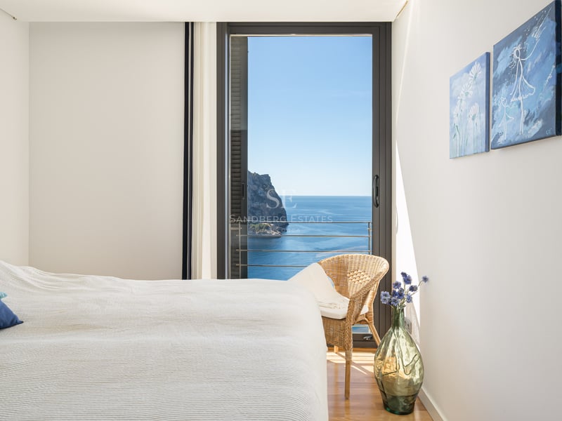 Bright bedroom with white linens, blue accent pillow, and floor-to-ceiling glass doors overlooking the Mediterranean sea.
