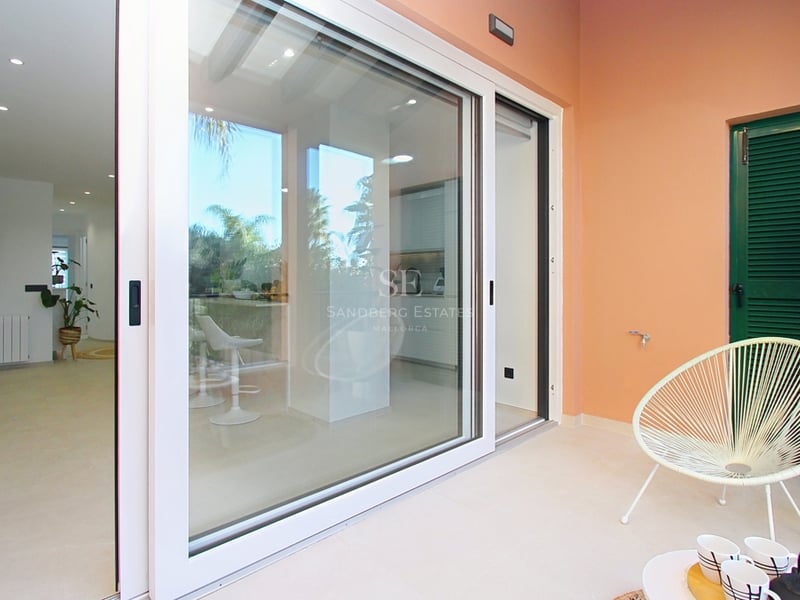 En solig terrass med en vit Acapulco-stol och skjutdörrar i glas som öppnar upp mot ett modernt vardagsrum.