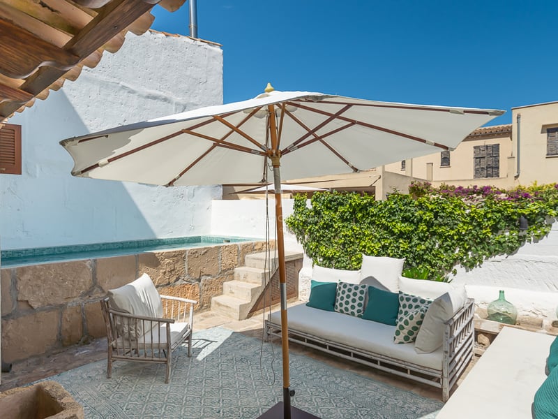 Patio méditerranéen ensoleillé avec piscine en pierre surélevée, canapé blanc et parasol.