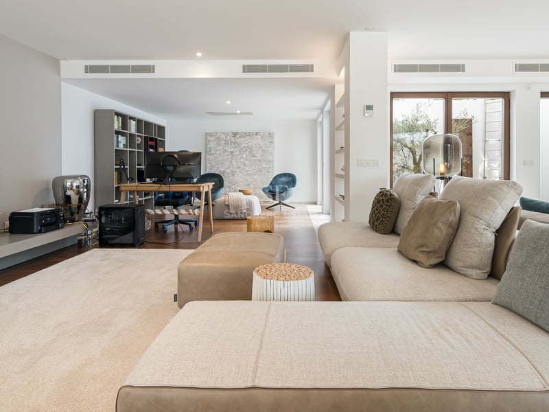 Salon contemporain avec grand canapé en L beige, parquet en bois et coin bureau au fond avec plusieurs écrans.