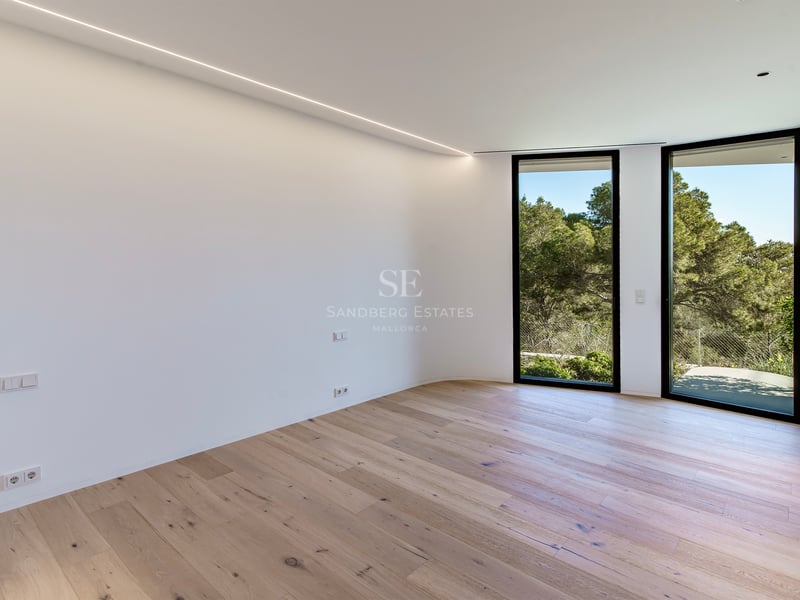 Pièce vide lumineuse avec parquet en bois clair, murs blancs et grandes fenêtres donnant sur la forêt.