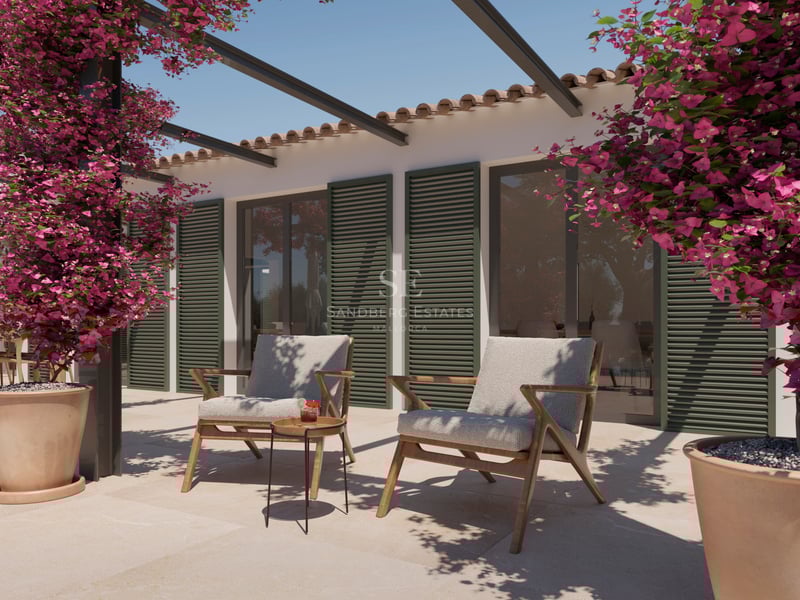 Terrasse en pierre avec deux fauteuils en bois, volets verts et bougainvilliers roses éclatants sous un ciel clair.