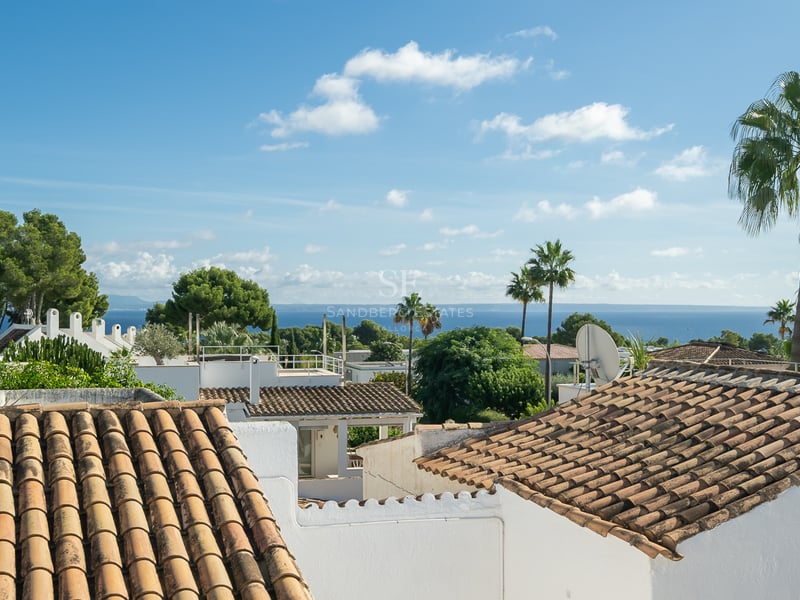 Vue surélevée de toits en terre cuite, de murs blancs et de palmiers avec la mer bleue en arrière-plan.