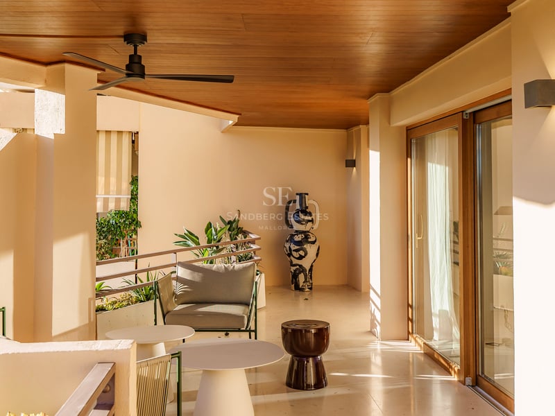 Überdachte Terrasse mit hellem Steinboden, modernen Möbeln, einem Deckenventilator und einer großen dekorativen Vase.