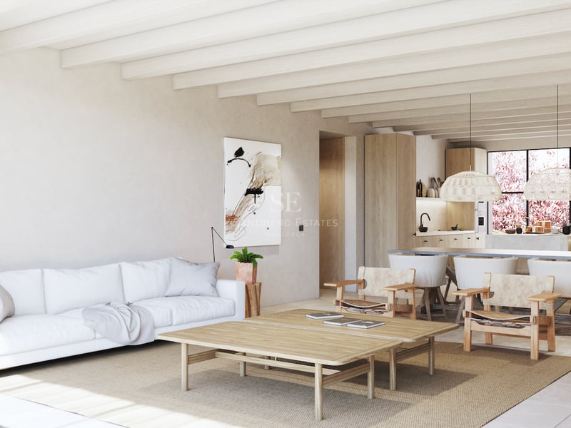 Salon lumineux avec poutres apparentes, canapé blanc, mobilier en bois clair et cuisine ouverte à l'arrière-plan.