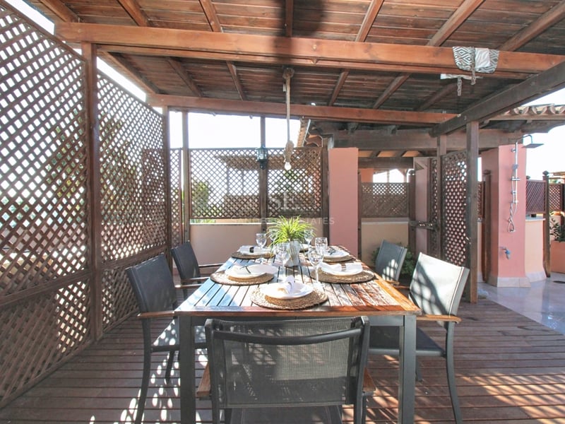 Matsalsbord under en träpergola med insynsskydd och trädäck på en solig terrass.