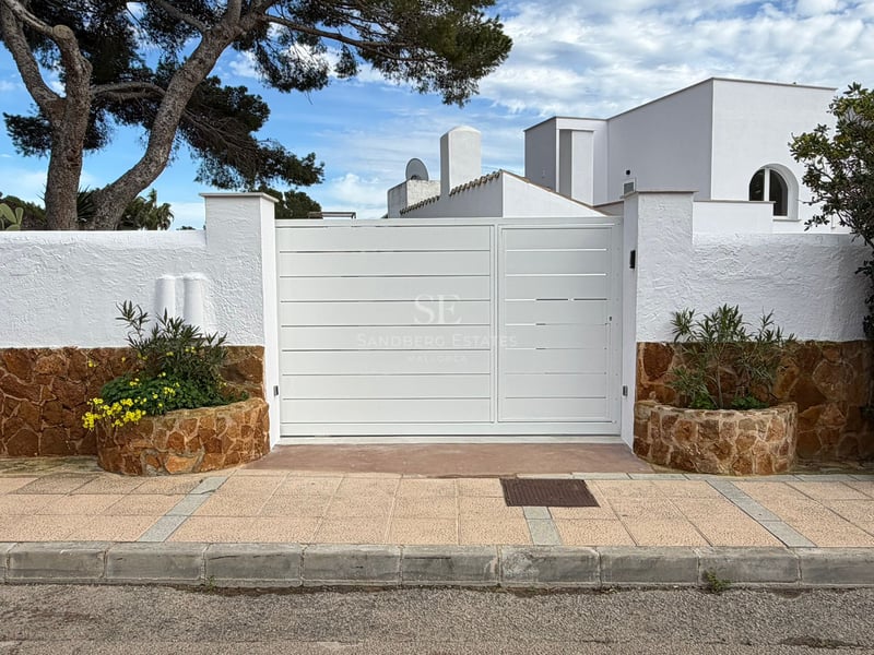 Grand portail métallique blanc flanqué de murs en stuc blanc et pierre marron sous un ciel bleu avec un pin.