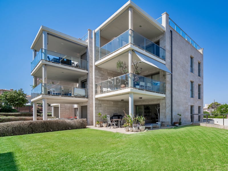 Villa contemporaine de trois étages avec revêtement en pierre, balustrades en verre et pelouse verte sous un ciel bleu.