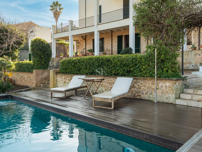 Turkos pool med mörkt trädäck, solstolar och rustik stenvägg i bakgrunden.