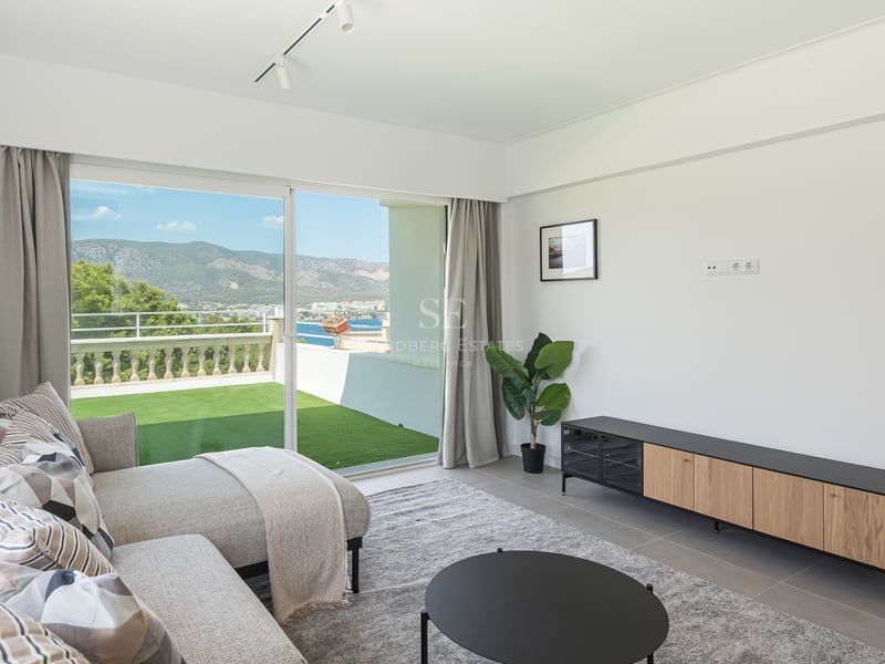 Salon moderne avec canapé gris et baies vitrées ouvrant sur une terrasse vue mer et montagne.