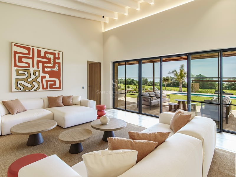 Heller Wohnraum mit weißen Sofas, Holztischen und bodentiefen Fenstern mit Blick auf Pool und Garten.