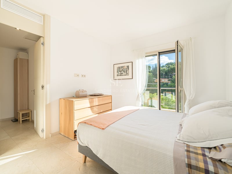 Ljust sovrum med säng, byrå i ljust trä och öppen balkong med utsikt över grönska.