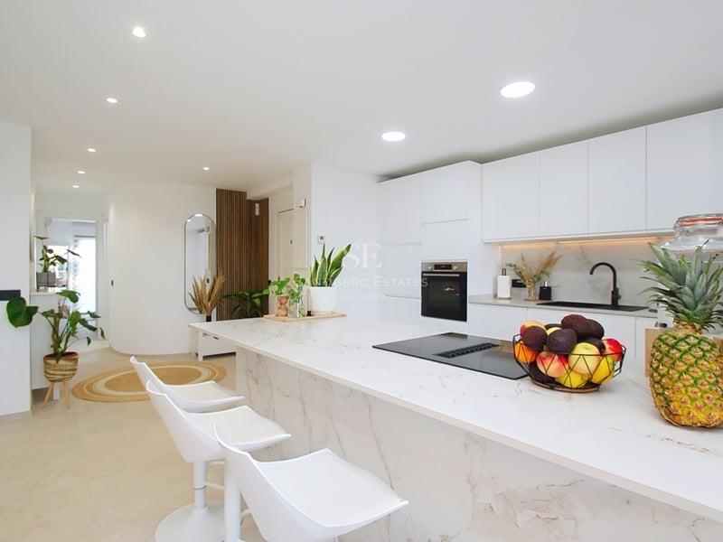 Minimalistiskt vitt kök med marmorliknande köksö, vita barstolar och inbyggda vitvaror.