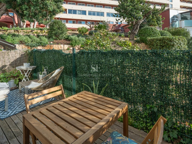 Sun-drenched private terrace featuring a wooden dining set, lounge area with patterned rug, and green privacy fencing.