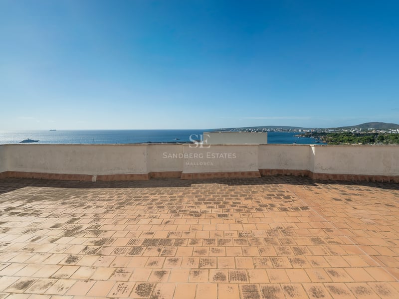 Large rooftop terrace with terracotta tiles overlooking a clear blue sea and coastline under a bright sky.