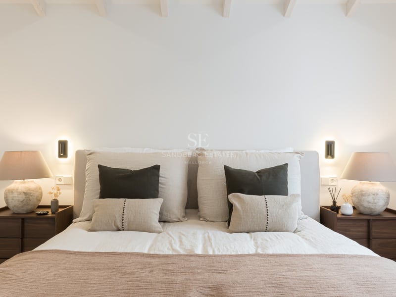 Lit avec tête de lit rembourrée, coussins décoratifs, tables de chevet en bois et lampes design à lumière chaude.