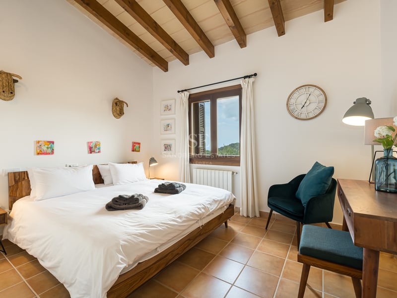 Chambre avec sols en terre cuite, poutres en bois apparentes, literie blanche et fauteuil en velours bleu canard.