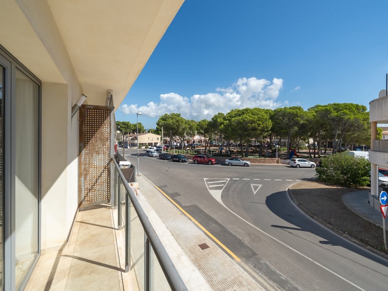 Modern gräddfärgad balkong med glasräcke med utsikt över en trädkantad gata och torg under en klarblå himmel.