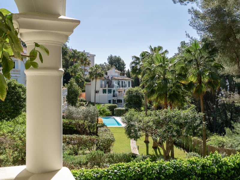 View from a white terrace column of a lush green garden with a blue swimming pool and palm trees under a clear sky.