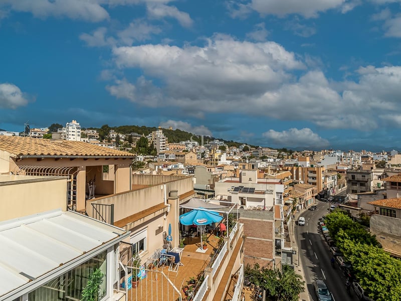 Erhöhte Ansicht mediterraner Hausdächer, einer von Bäumen gesäumten Straße und ferner Berge unter blauem Himmel.