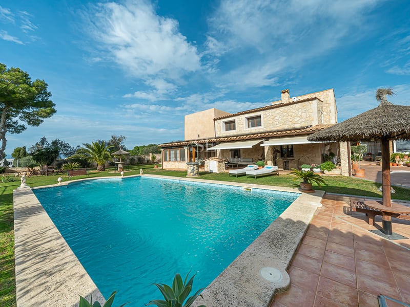 Grande piscine turquoise devant une villa en pierre avec jardin soigné, pelouse et parasols en chaume.