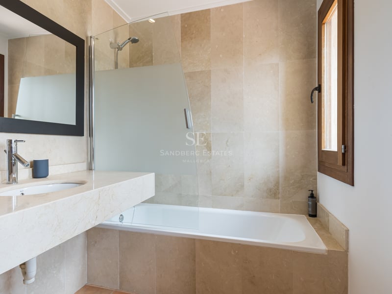 Salle de bain moderne avec carrelage en pierre beige, baignoire blanche, paroi en verre et fenêtre en bois.