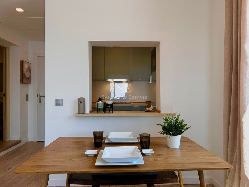 Wooden dining table set for two in front of a wall opening looking into a modern kitchen.